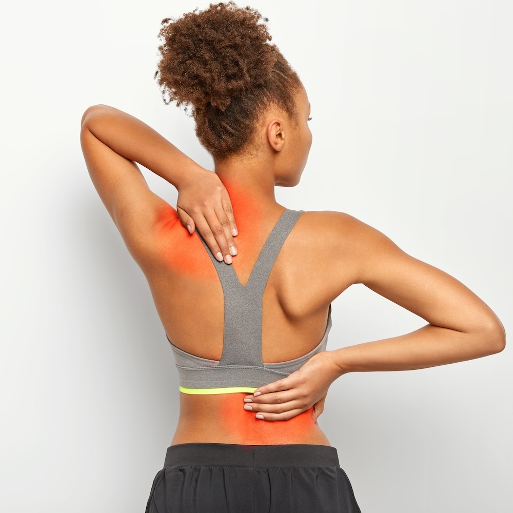 Faceless curly woman suffers from spine pain, has injury after overtraining, wears sport bra, shows location of inflammation, leads sporty lifestyle, isolated on white background. Chronic disease